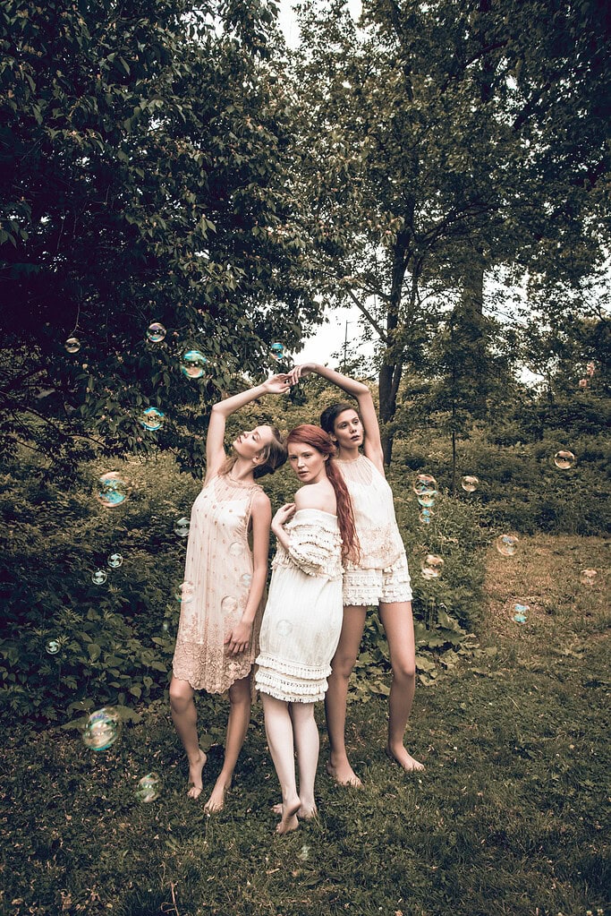 Three fashion models in pale dresses posing beneath raised arms with bubbles floating around them.