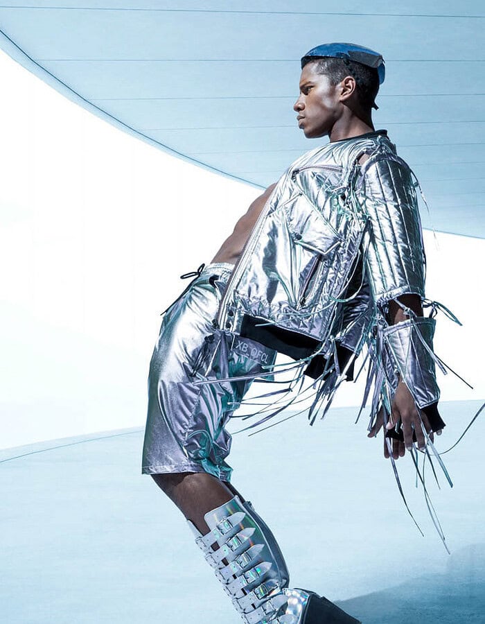 Male fashion model in a silver outfit and platform boots leaning across a cool blue futuristic set.