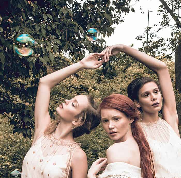 Three fashion models in pale dresses posing beneath raised arms with bubbles floating around them.