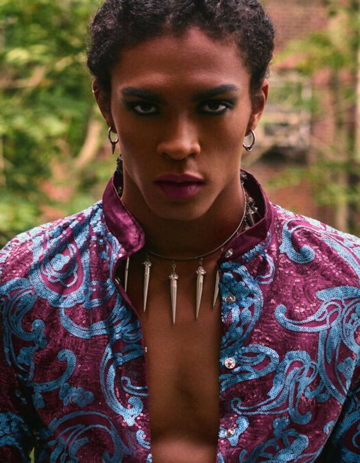 Close portrait of a menswear model wearing a burgundy and blue sequined jacket in a green courtyard.