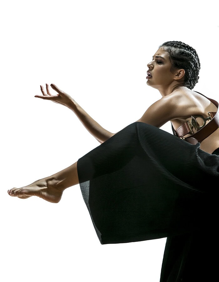 Fashion model in black pleated trousers posing with one leg extended and one hand raised.